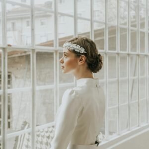 Pensive young female in white elegant dress and hairband standing near windows in room in daylight
